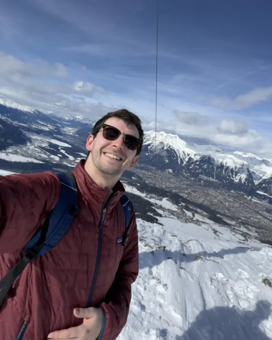 Sean McMahon, founder of Woerka, smiling on a snowy summit in the Alps, with mountains and a valley town behind